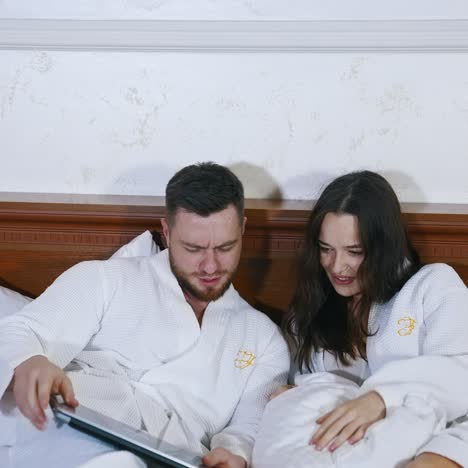 Happy couple rest in hotel. Woman and man in dressing gowns on bed. Married couple in white coats sit comfortably on bed to watch video on a laptop