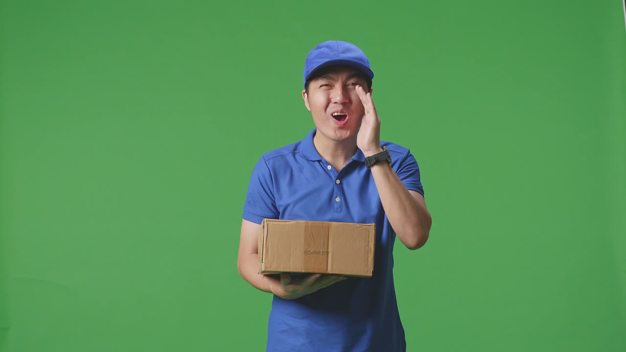 푸른 유니폼을 입은 아시아 남성 택배기사가 미소 짓고 스튜디오에서 녹색 스크린 배경에 상자를 배달하는 동안 카메라와 이야기합니다.