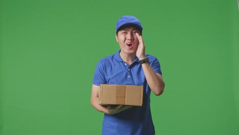 mensajero masculino asiático en uniforme azul sonriendo y hablando a la cámara mientras entrega una caja de cartón en el fondo de la pantalla verde en el estudio