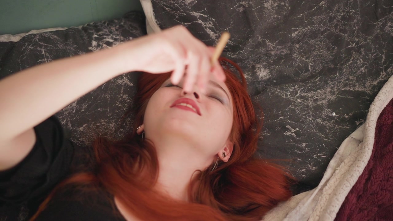 Caucasian redhead lying on bed smiling, holding slender stick, relaxed gaze, lighthearted expression, cozy textiles, imaginative mood, future oriented daydream, casual home portrait