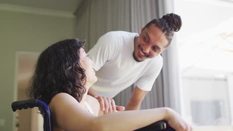 mujer biracial feliz en silla de ruedas y compañero masculino sonriente con rastas hablando en casa