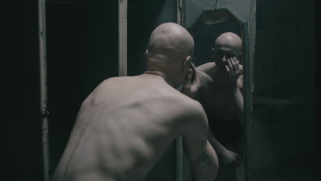 pale bald skinny man looks into the mirror in the creepy old bathroom at night