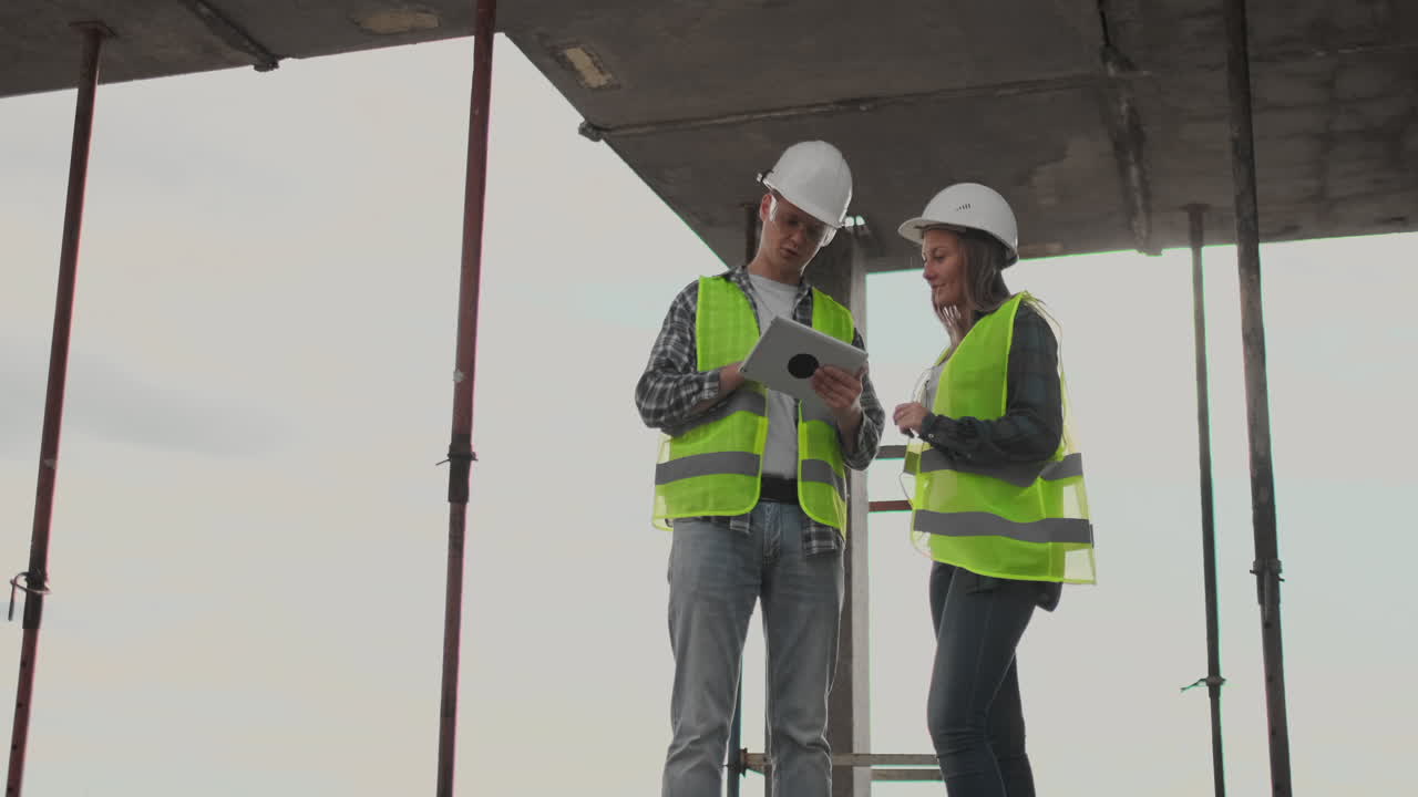 dos ingenieros un hombre y una mujer con cascos blancos con una tableta en el sitio de construcción condenan el plan de construcción del edificio