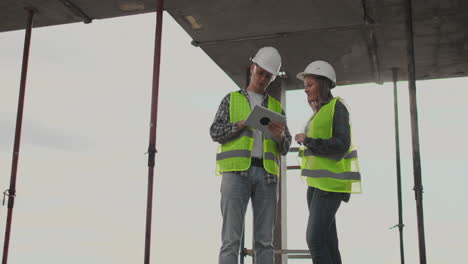 Dos-Ingenieros,-Un-Hombre-Y-Una-Mujer-Con-Cascos-Blancos-Y-Una-Tableta-En-El-Sitio-De-Construcción,-Condenan-El-Plan-De-Construcción-Del-Edificio