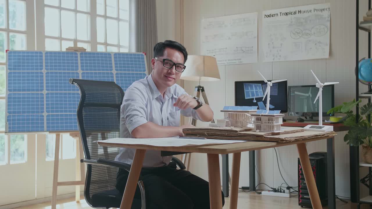 아시아인 남자가 태양 전지판을 가진 작은 집의 블루프린트와 모델로 작업하면서 카메라에 미소를 짓고 있습니다.