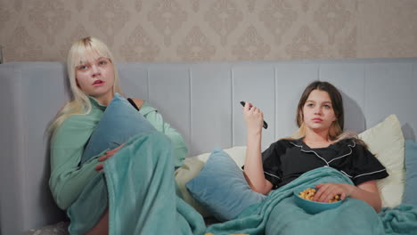 two teenage girls in pajamas relaxing on bed watching movie with bored expressions, one girl holds remote control while other hugs pillow, popcorn bowl sits on blanket