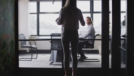 young white woman joins male creative in a meeting room for a job interview, seen through glass wall