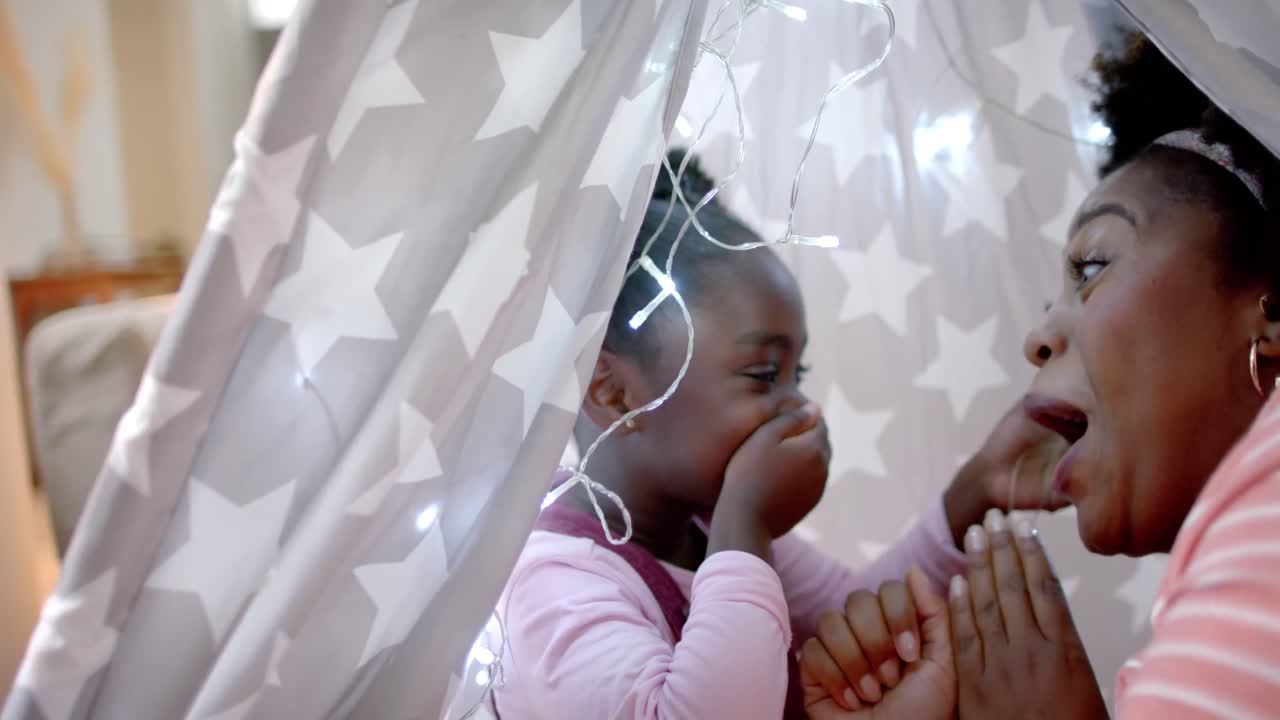 Happy african american mother and daughter laughing in tent at home, slow motion