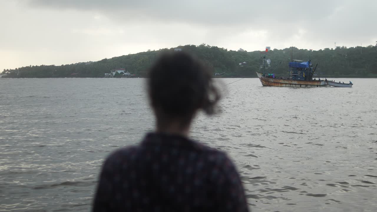 Woman blending with landscape, watching fishing boat gliding along Mandovi River under cloudy sky, embodying peaceful introspection in Goa, India