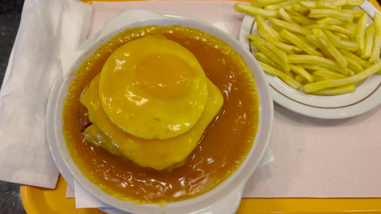 A hearty plate of Francesinha, a Portuguese sandwich filled with layers of meat and cheese, covered in a savory sauce and topped with a fried egg. Served alongside crispy fries for a filling meal.
