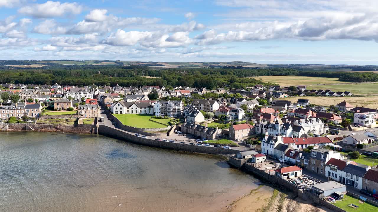 Drone glides toward shoreline village, revealing houses, countryside, and bay under bright daylight