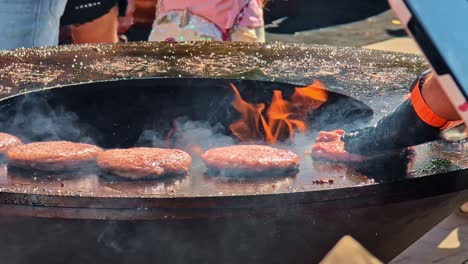 A close-up shot shows a chef grilling juicy beef burger patties on a large, round fire pit grill, with flames and smoke rising from the open fire at an outdoor food festival