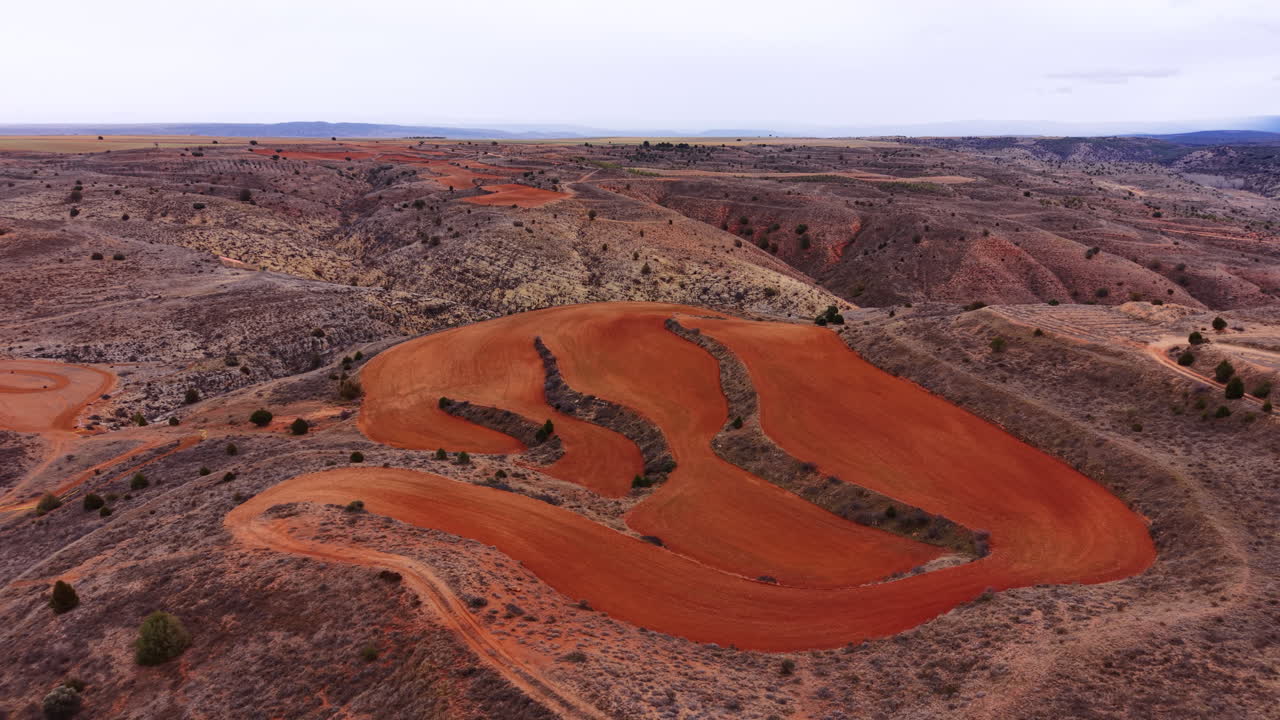 Campo agricolo a terrazze con terreno rosso vivido su un versante collinare eroso in Aragona, Spagna, circondato da un paesaggio di calanchi aridi.
