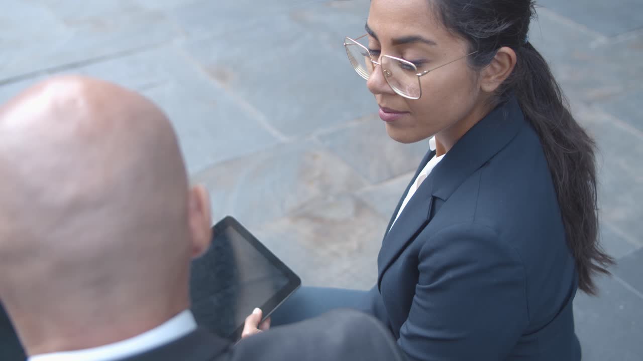 Latin businesswoman listening to male colleague, holding tablet while sitting outside