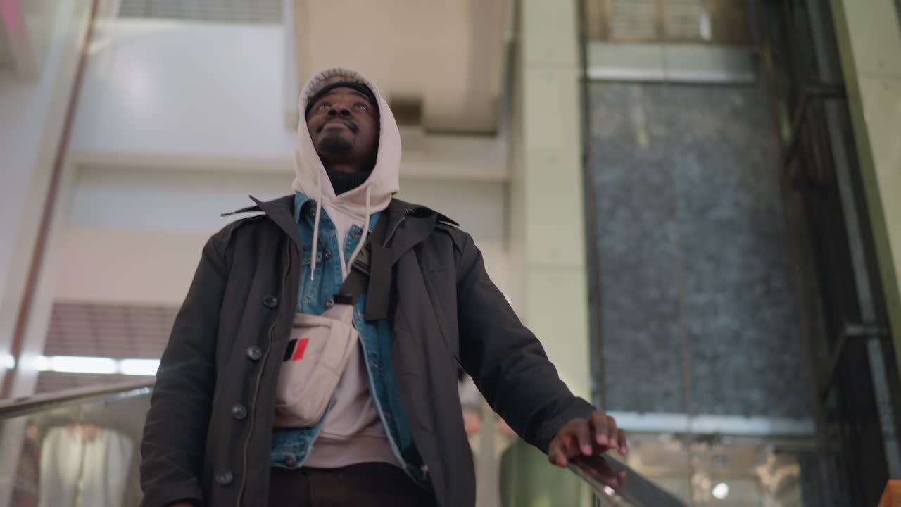 Young man in hoodie and jacket standing on escalator in modern shopping mall, focused expression, casual outfit with small bag, soft lighting highlighting his features