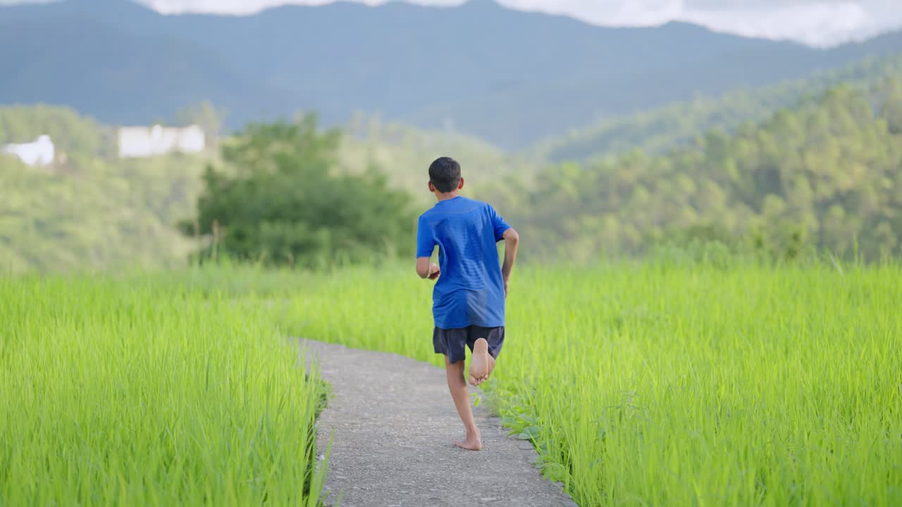 Indian boy in blue T-shirt running barefoot across flooded paddy fields, joyful rural childhood scene, 4k video