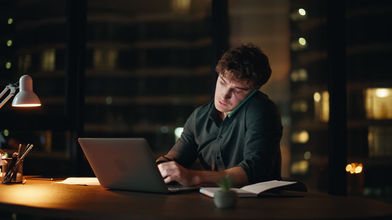 Freelancer sovraccarico di lavoro che parla al cellulare e guarda il portatile in ufficio a tarda sera