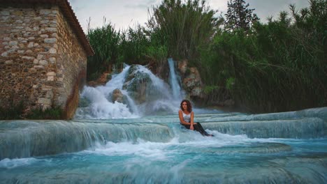 cinemagraph de 4k uhd: bucle de video continuo de una mujer joven sentada frente a una cascada de aguas termales en saturnia, italia