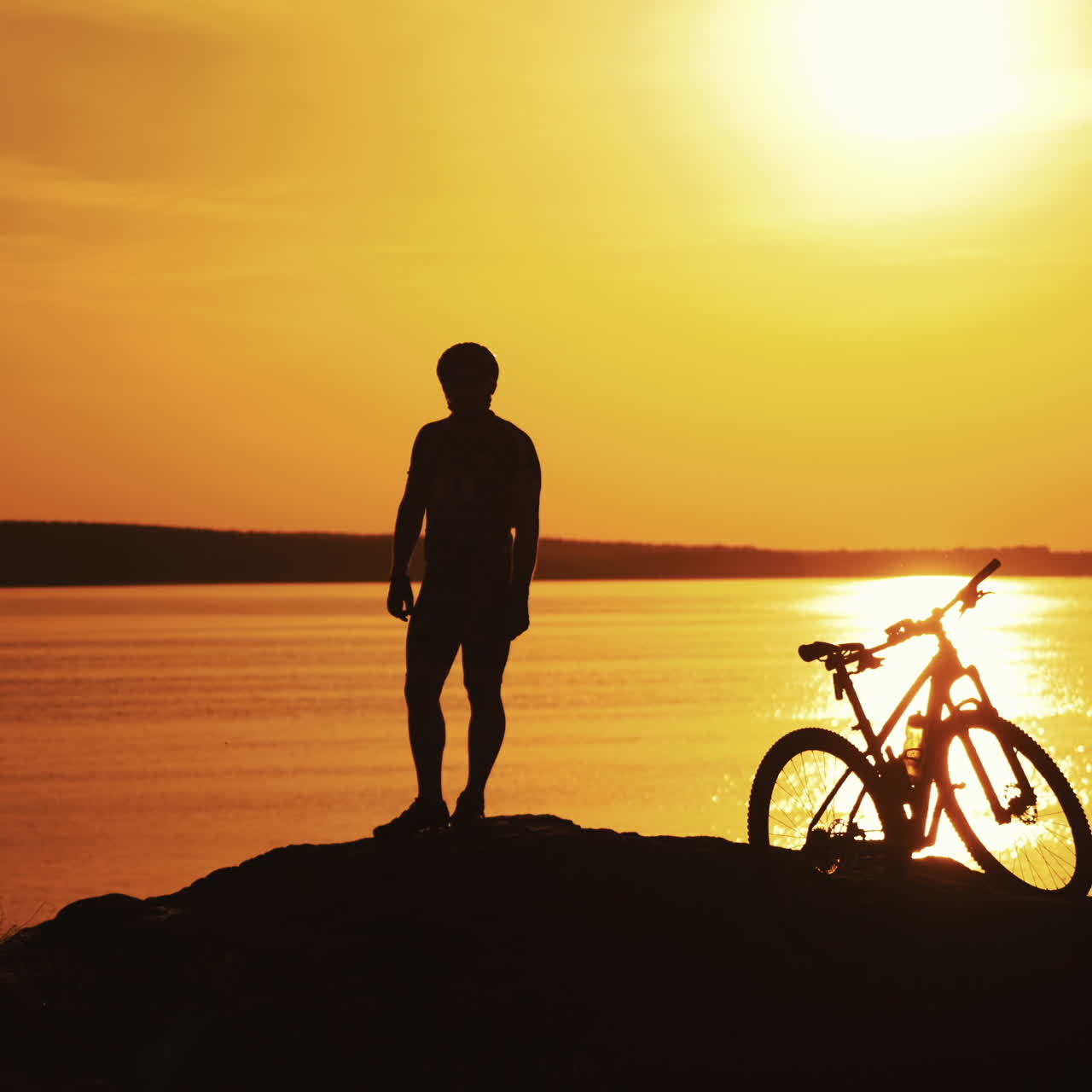 Male bike rider is standing next to his bike near the river at sunset. Silhouette of a biker in helmet who is having a rest on the orange water background. Evening landscape and a cyclist.