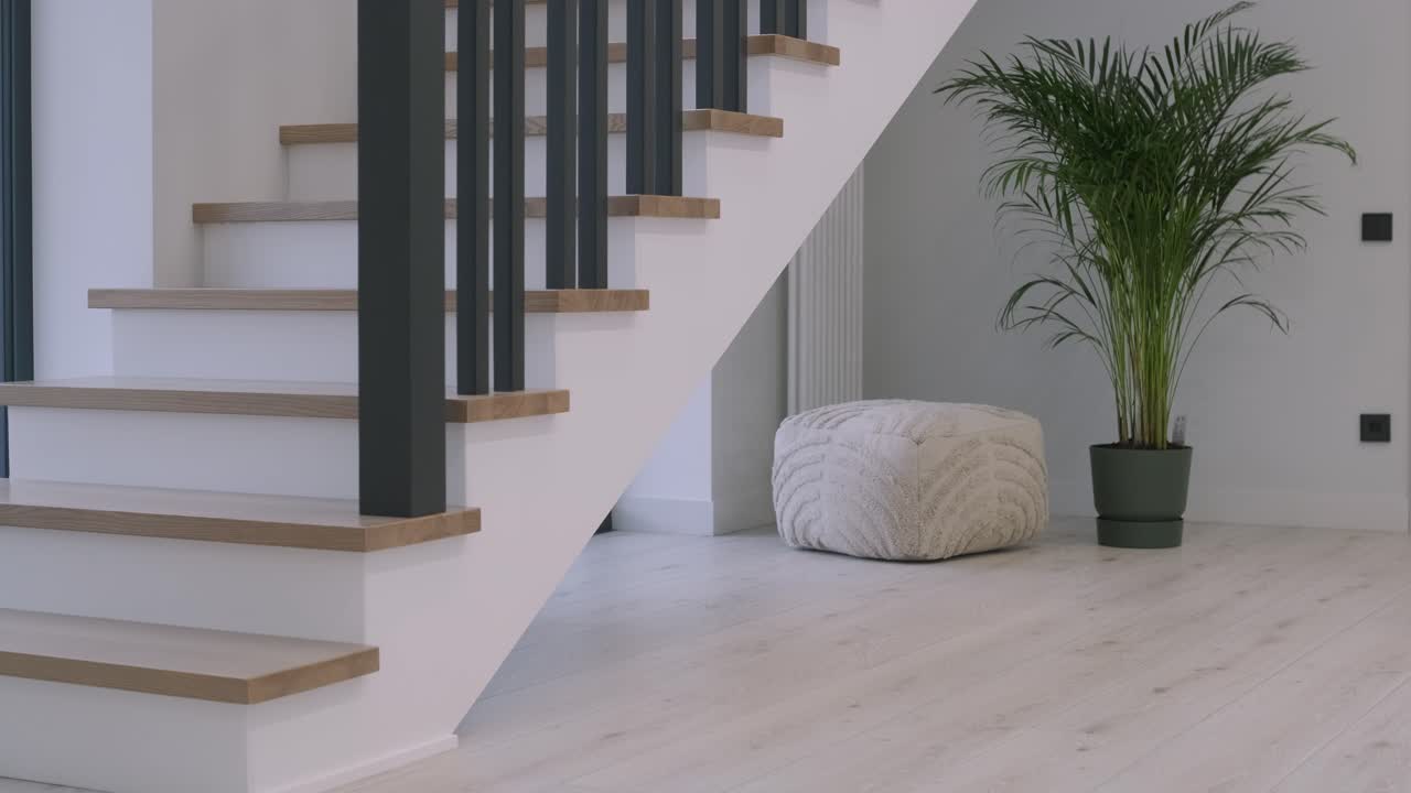 A part of a modern home interior with a stylish staircase featuring wooden steps and black vertical balusters, a cozy pouf on the floor, and a tall green potted plant enhancing the space with a touch of nature