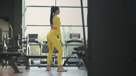 young hispanic brunette woman squats holding a dumbbell in the gym in a yellow tracksuit