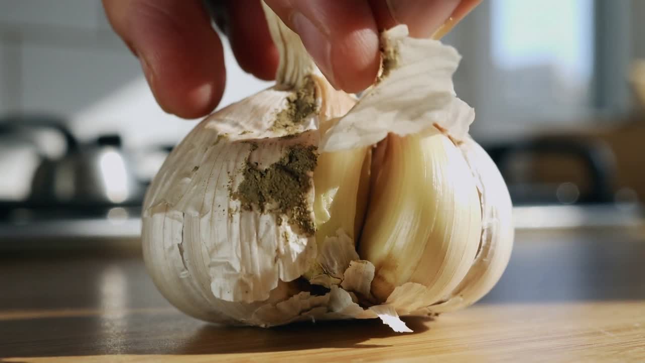 Erwachsene Finger halten eine Knoblauchknolle fest, ziehen auf einem Küchenbrett die papierartige Haut ab und legen die Zehen frei.