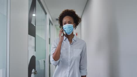 Mixed-race-businesswoman-in-face-mask-using-smartphone-walking-in-corridor-in-office