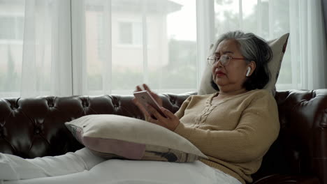 집에서 휴식을 취하는 활동적인 고령 여성 블루투스 이어폰을 착용하여 소파에 편안하게 앉아 휴대 전화로 음악을 듣는 것을 즐기십시오.