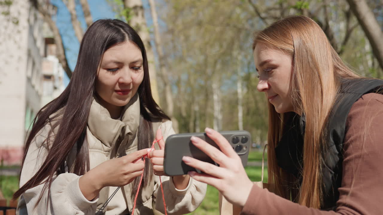 Unas amigas examinan un pequeño dispositivo junto a un teléfono; una mujer asiática y otra de raza blanca analizan el embalaje de unos auriculares con atención; primer plano de los dedos y el dispositivo; expresiones de curiosidad y ambiente de desempaquetado en un parque soleado.