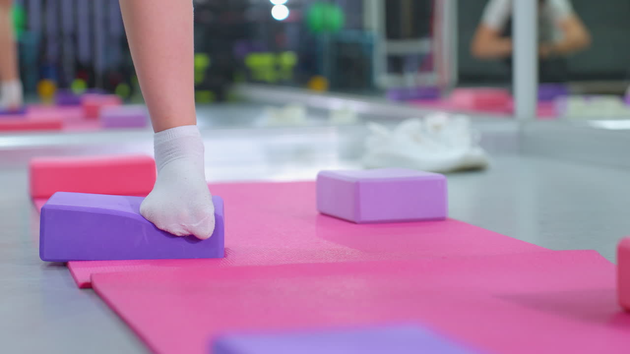 색 양말을 입고 보라색 요가 블록에서 균형을 잡고 있는 사람의 다리 시각 핑크색 운동 매트에서 안정 훈련을 하는 동안, 체육관 환경에서 핵심 힘, 조율 및 균형 개선에 초점을 맞추고 있습니다.