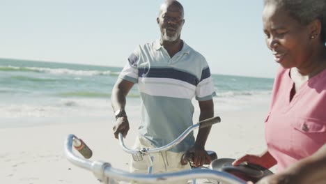 Happy-senior-african-american-couple-walking-with-bikes-at-beach,-in-slow-motion