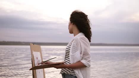Outstanding view of a young curly brunette artist in working process, standing in the water with easel and drawing a picture with surrounding landscape. Outdoors