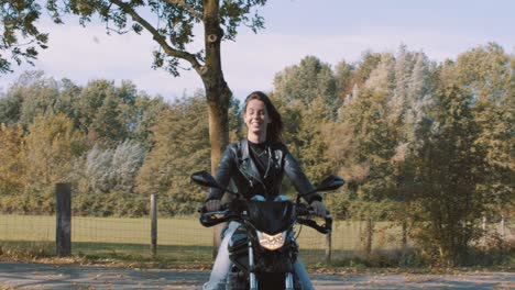 pretty smiling european young woman driving a motorbike wearing leather jacket in forest with vibrant, colorful golden autumn leaves on sunny day