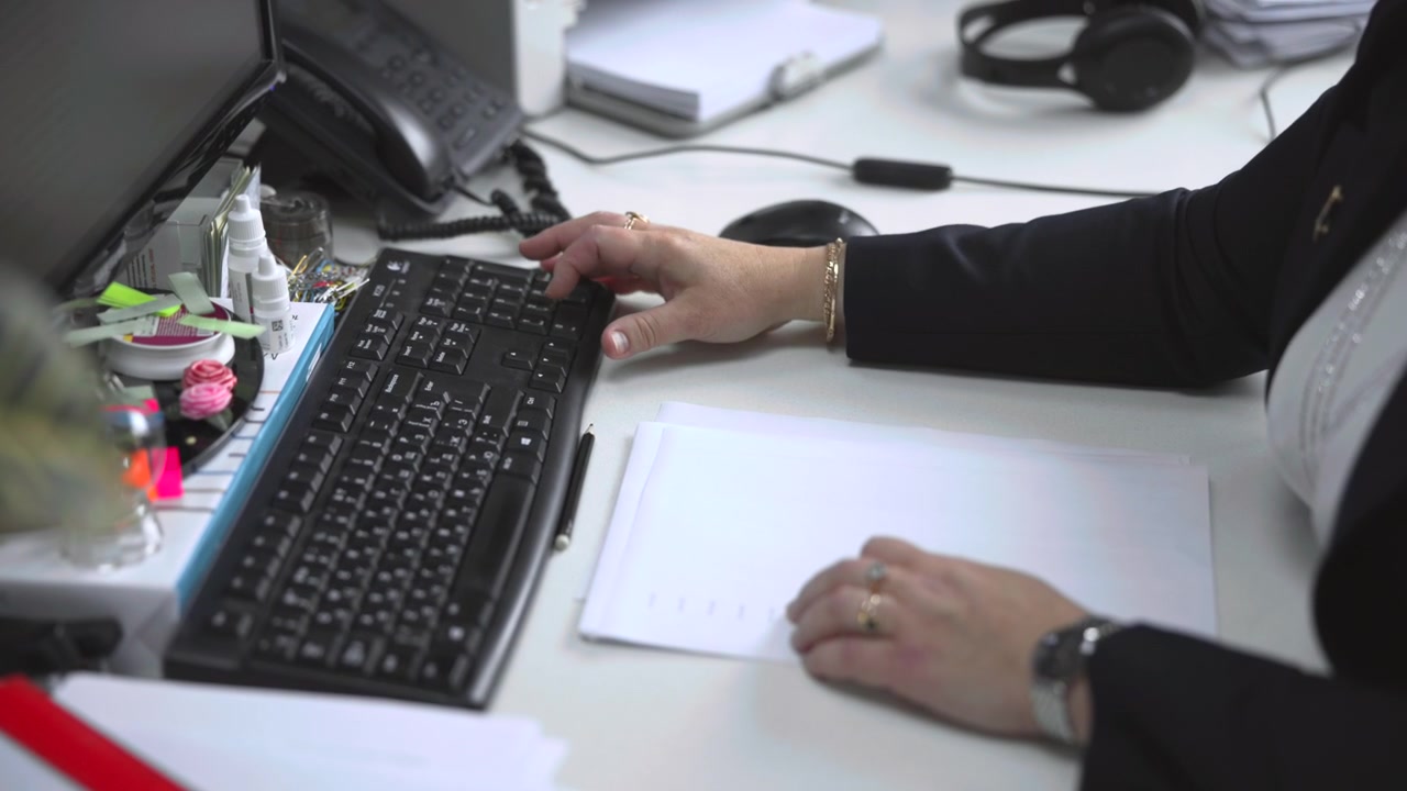 Eye level close view showing office worker hands typing on full size black keyboard while other hand steadies stack of white documents on pale desk with mouse, telephone, headset, and office supplies
