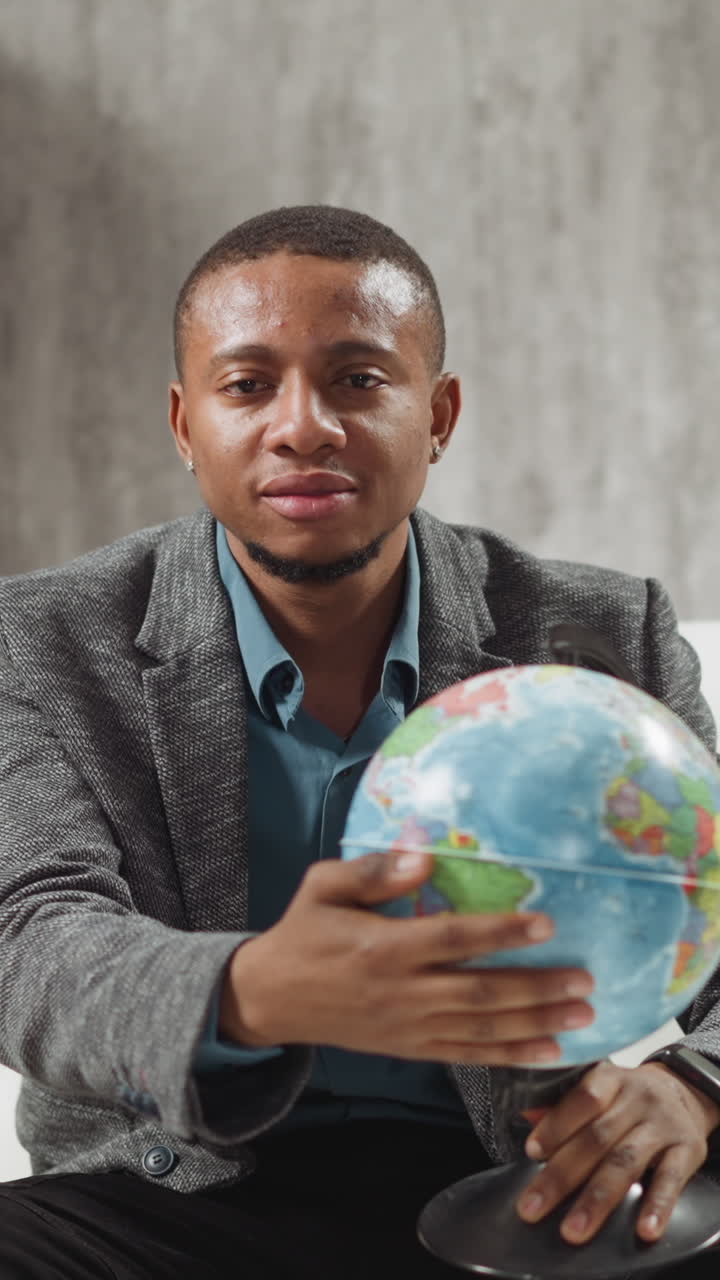 Positive African-American geography teacher turns globe inviting students to online lesson sitting near laptop in private apartment slow motion