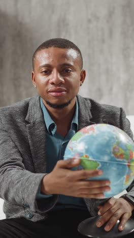 positive african-american geography teacher turns globe inviting students to online lesson sitting near laptop in private apartment slow motion