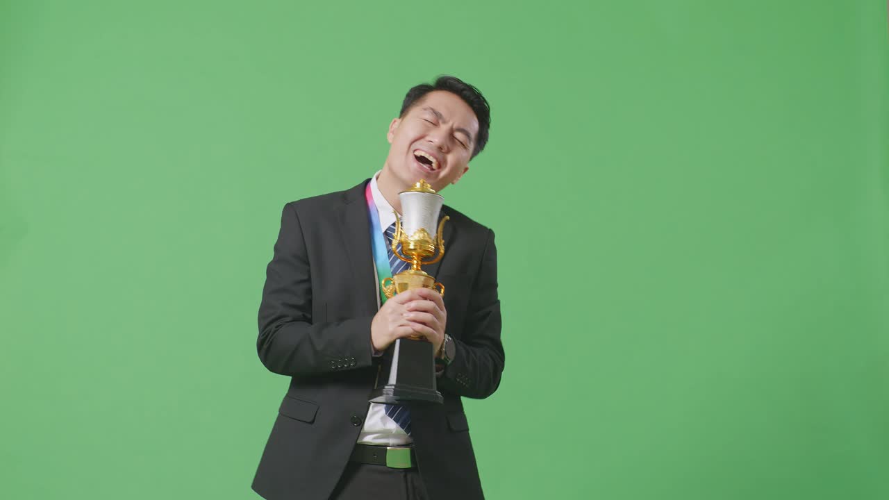 슈트와 타이를 입은 아시아 사업가, 금메달과 함께 금 트로피에서 노래하고, 스튜디오의 녹색 스크린 배경에서 첫 번째 우승자로 우승을 축하합니다.