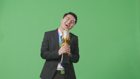 hombre de negocios asiático en traje y corbata con una medalla de oro cantando en un trofeo de oro celebrando la victoria como el primer ganador en el fondo de la pantalla verde en el estudio