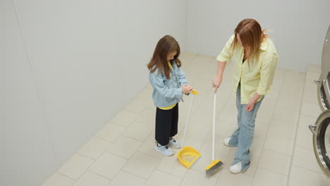 Aerial view shows cheerful mother sweeping tiled floor while toddler holds dustpan, both engaged in household cleaning, smiling and discussing while working together with broom and dustpan