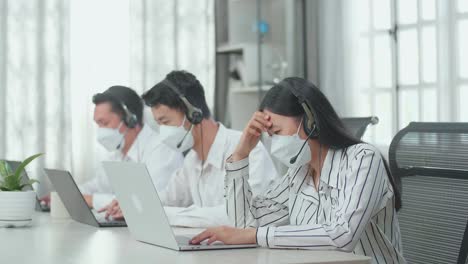 a woman of three asian call center agents wearing headset and mask headache to work while two of her colleagues are speaking and typing during the call with customers at the office