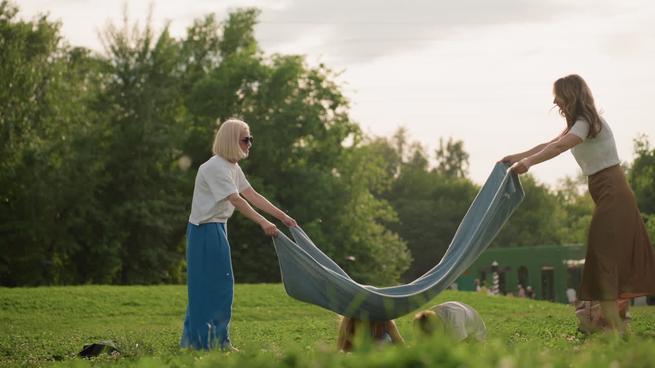 Nurturing family women and kids in park playing, swinging cloth over children, laughter and motion, green grass background, warm daylight casting soft glow, casual summer clothing