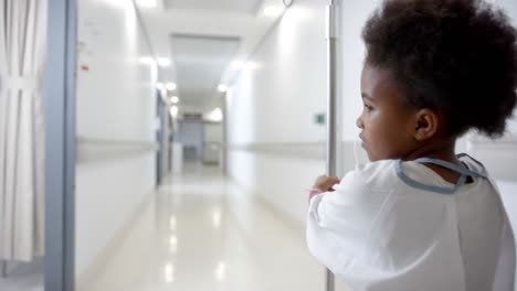 back of african american girl in hospital gown with drip walking in hospital corridor, slow motion
