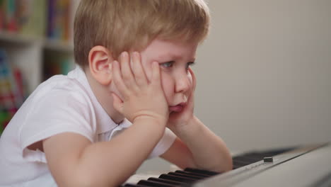 Little-boy-puts-elbows-on-piano-keyboard-and-leans-on-hands
