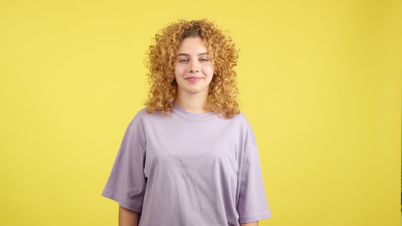 Young woman with curly blonde hair smiles and gives a thumbs up on a yellow background