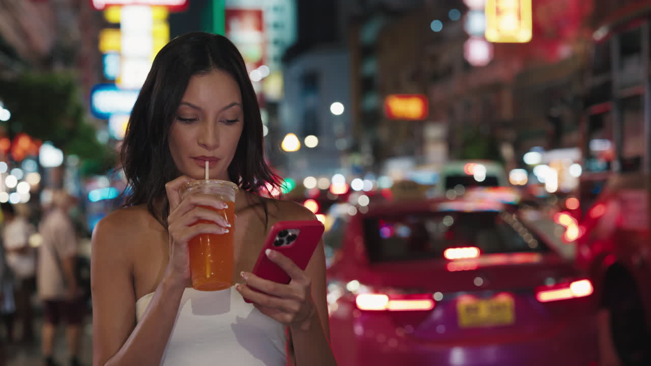 Woman enjoying a drink and using her phone on a bustling night street