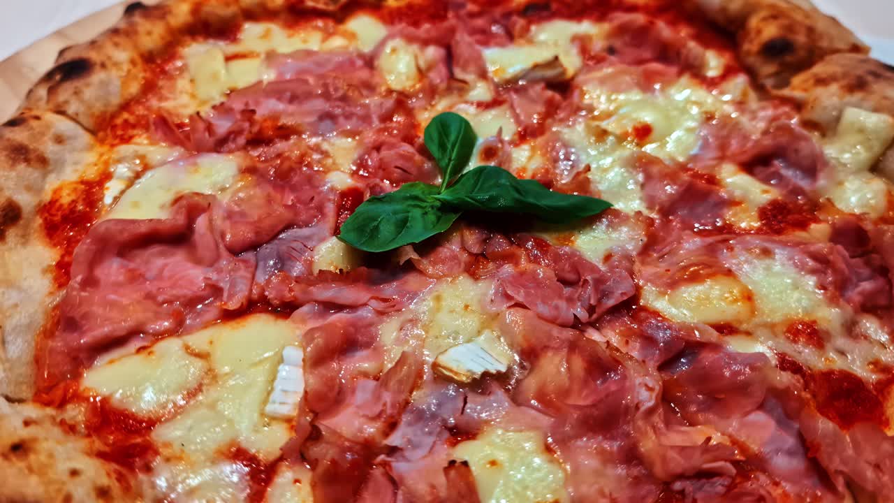 Close-up of a delicious and fresh ham and cheese pizza with basil leaves and a thick baked crust - parallax shot