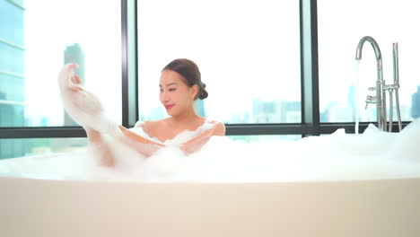 asian young woman playing with foam in the bathtub at high floor apartment with glass wall and city view, girl is spreading foam on her body and arms