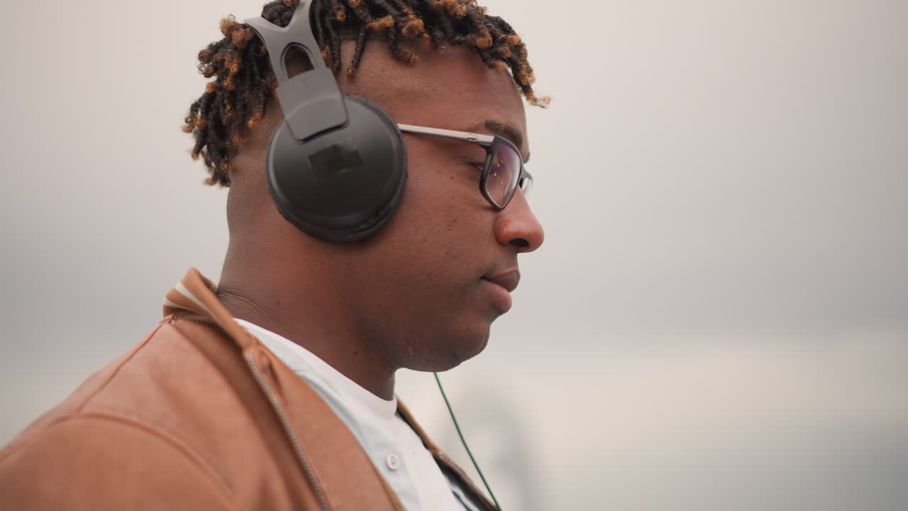 Svart mannlig utendørs DJ justerer hodetelefonene, fokusert profil med briller og brun jakke under overskyet himmel. Lydsjekk og subtil miksebevegelse, lo-fi urban stemning, nærbilde som fanger konsentrasjon.