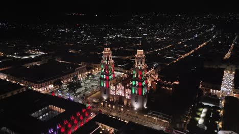 morelia cathedral at night, lit with the patriotic colors green, white, and red. drone shot during the month of september.