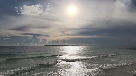 Backwards-flying-low-hanging-drone-over-calm-waves-coming-ashore-in-direct-sunlight